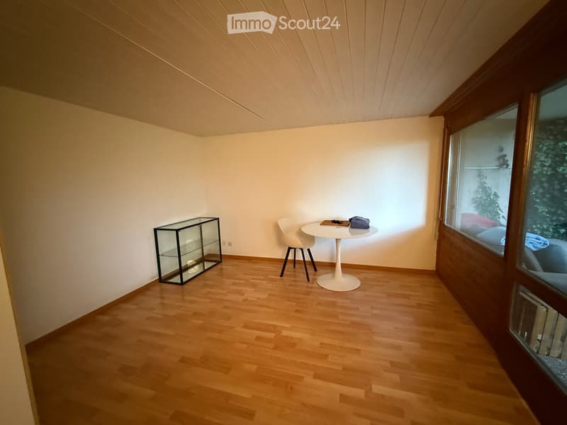 Bright, minimalist room with wooden flooring, white walls, and large windows. Includes a white round table with a chair, a glass display cabinet, and a small shelf with items visible through the window.