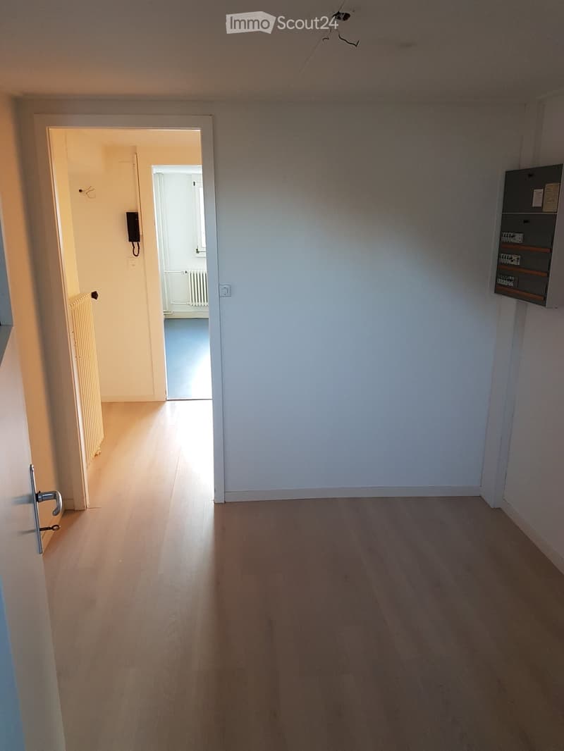 An empty room with a wooden floor, white walls, open door, electrical panel, and another room visible at the end