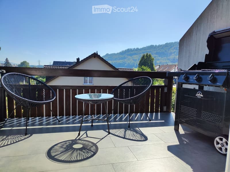 Balcony with two black wicker chairs, a small glass table, and a grill, overlooking a scenic landscape.