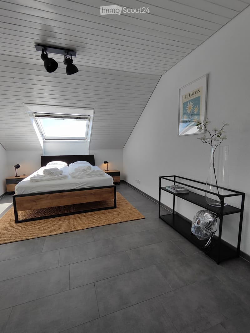 Modern bedroom with a wooden bed frame, nightstand, a sleek black table with a vase and fan, and a large window with blinds.
