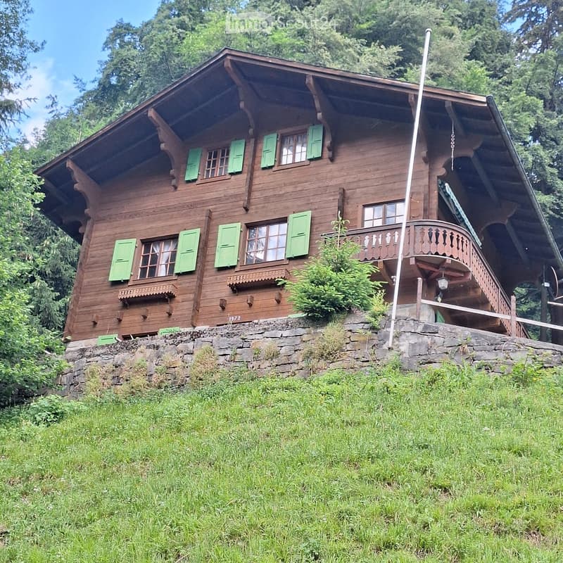 A two-story wooden house with green shutters, a stone foundation, and a balcony on the upper level, surrounded by greenery.