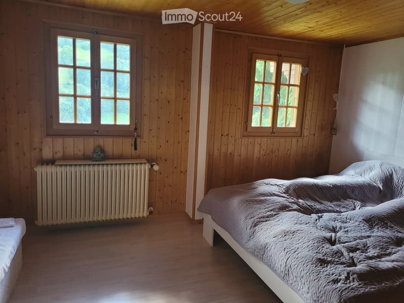Cozy bedroom with wooden walls and ceiling, large windows, a double bed with grey bedding, and a radiator.