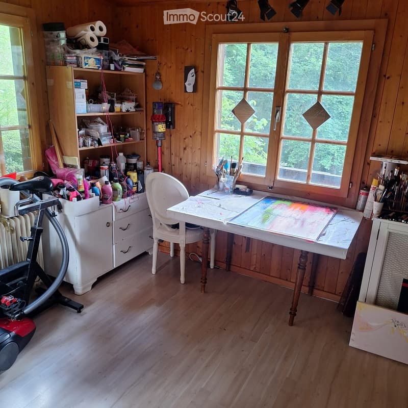 A well-lit studio with wooden walls and floors, featuring a white wooden chair, a table with paintings, shelves filled with art supplies, and an exercise bike in the corner.