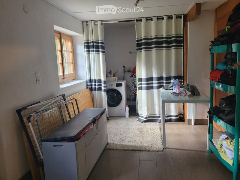 Laundry room with a washing machine, ironing board, small table, and shelf with folded clothes.
