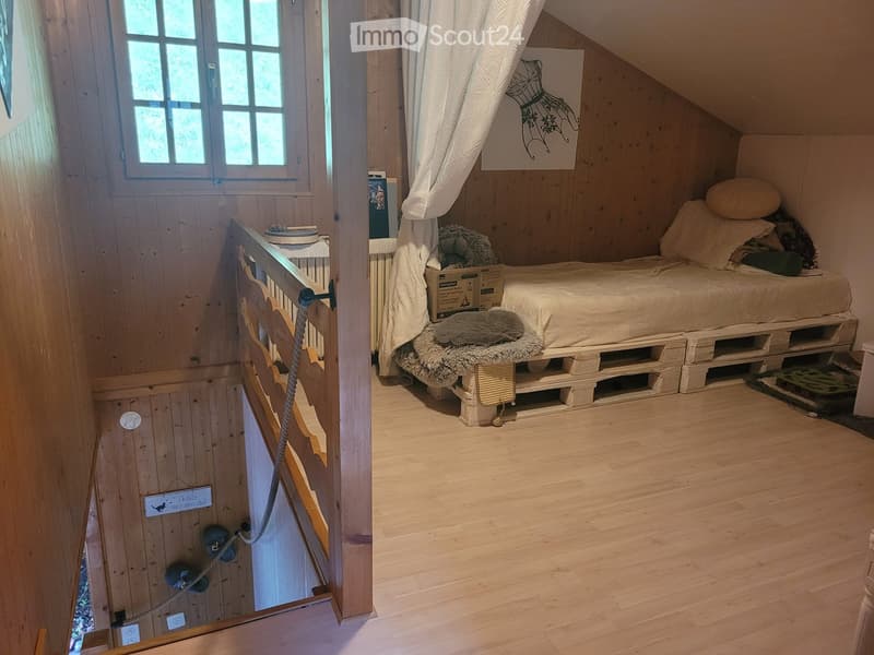 Cozy attic bedroom with wooden walls and floor, featuring a bed made from wooden pallets, pillows, a blanket, and a radiator. A curtain and picture frame are visible on the wall.