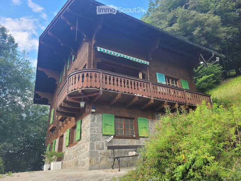 A two-story wooden house with green shutters, a balcony, and stone base, surrounded by greenery and a bench.