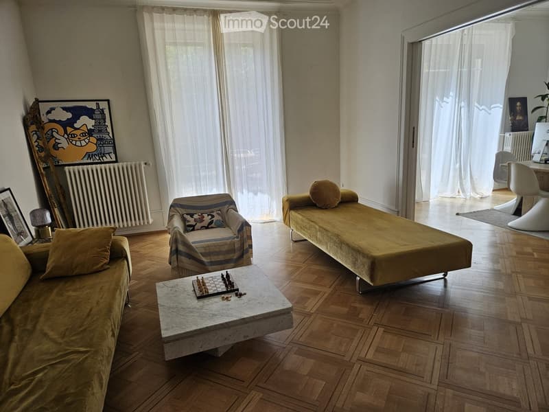 Living room with wooden flooring, large windows with white curtains, a sofa, a chair, a coffee table with a chessboard, and a small table with chairs.