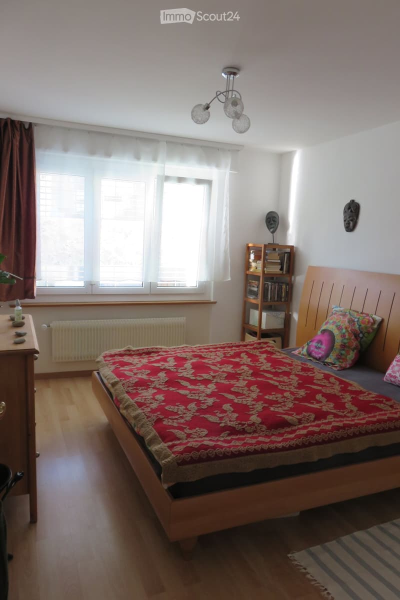 A well-lit bedroom with wooden flooring, a large bed with a red and gold patterned bedspread, colorful pillows, a wooden bookshelf, a small table with a bottle, and decorative masks on the wall.