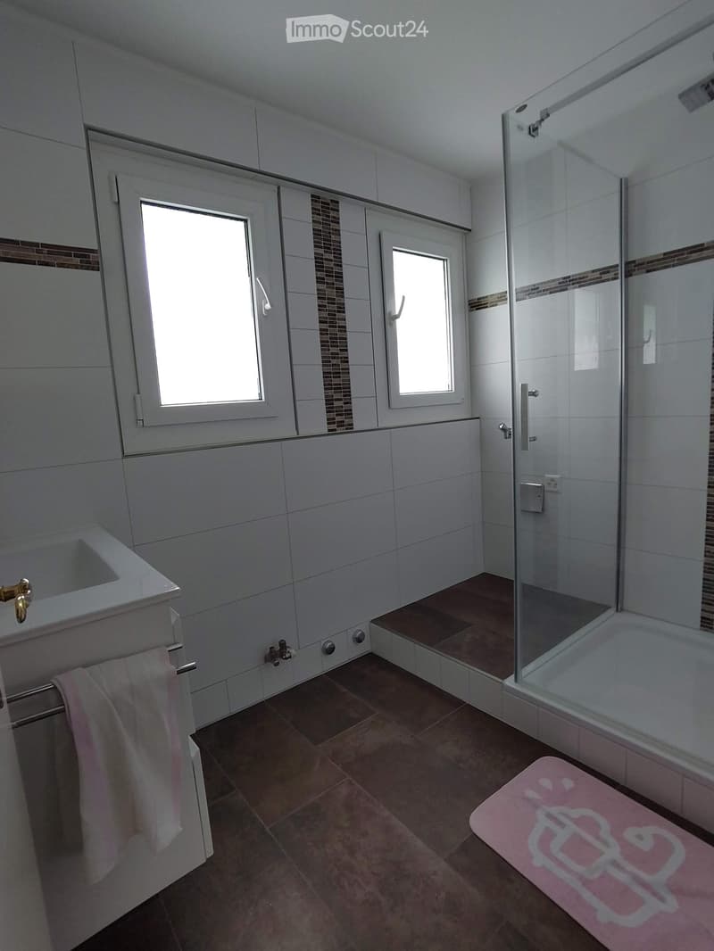 Modern bathroom with white tiles, a bathtub, a glass-enclosed shower, a sink with a mirror, and a pink bath mat with a cartoon character design.