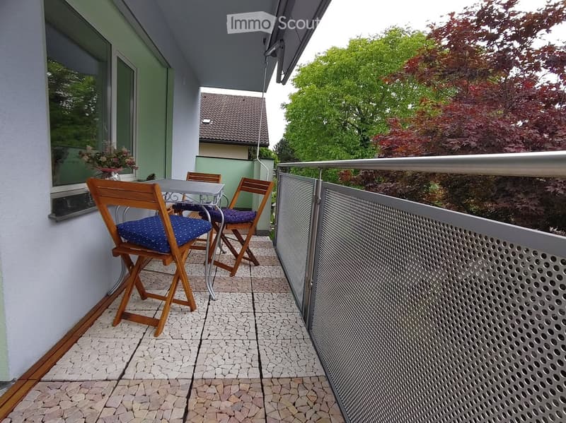 Balcony with a small table and two wooden chairs, featuring a metal railing and a view of greenery.