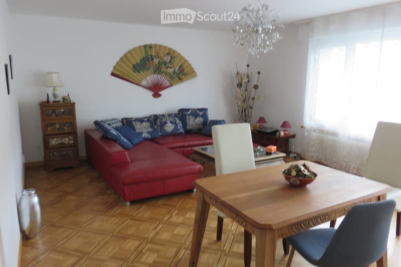 A well-lit living room with a red leather sofa, wooden dining table, chairs, decorative fan, and lamps.