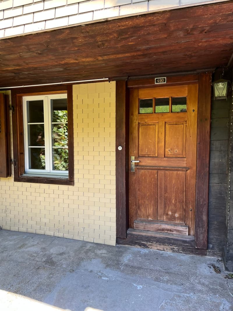 Front entrance of a house with a wooden door, a window to the left, and a small porch area.