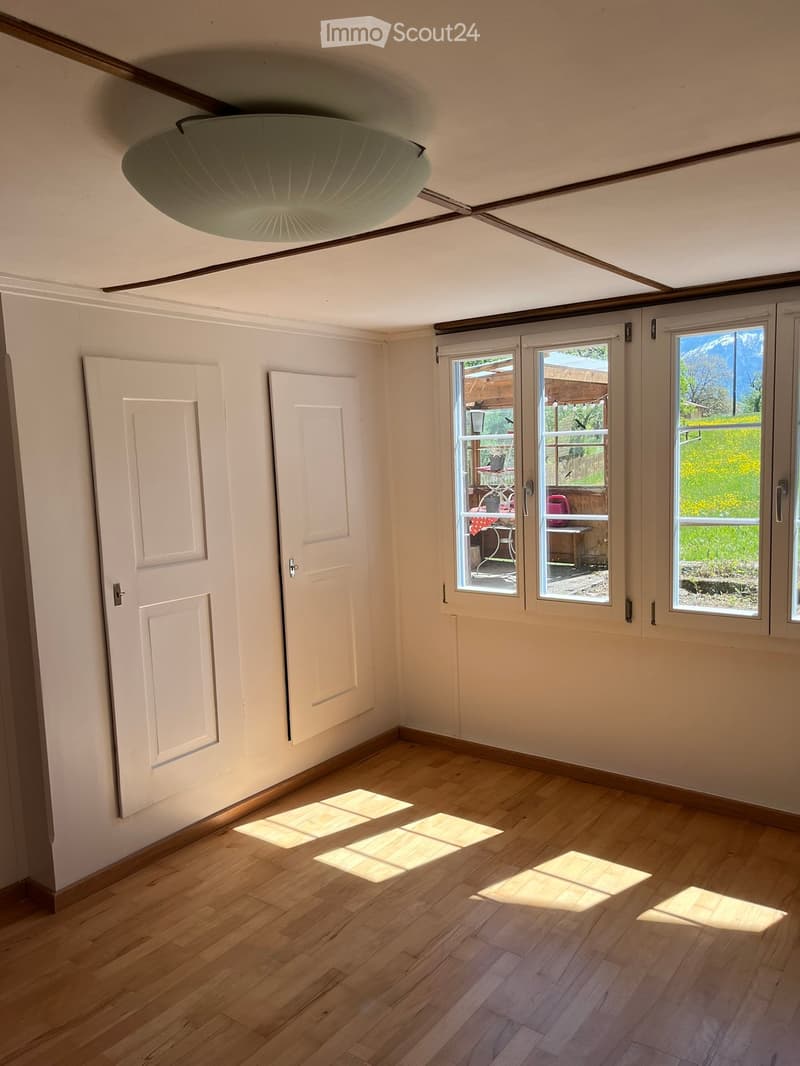 Empty room with wooden flooring, white walls, and large windows offering a view of the outdoors. Two white doors are visible on the left side.