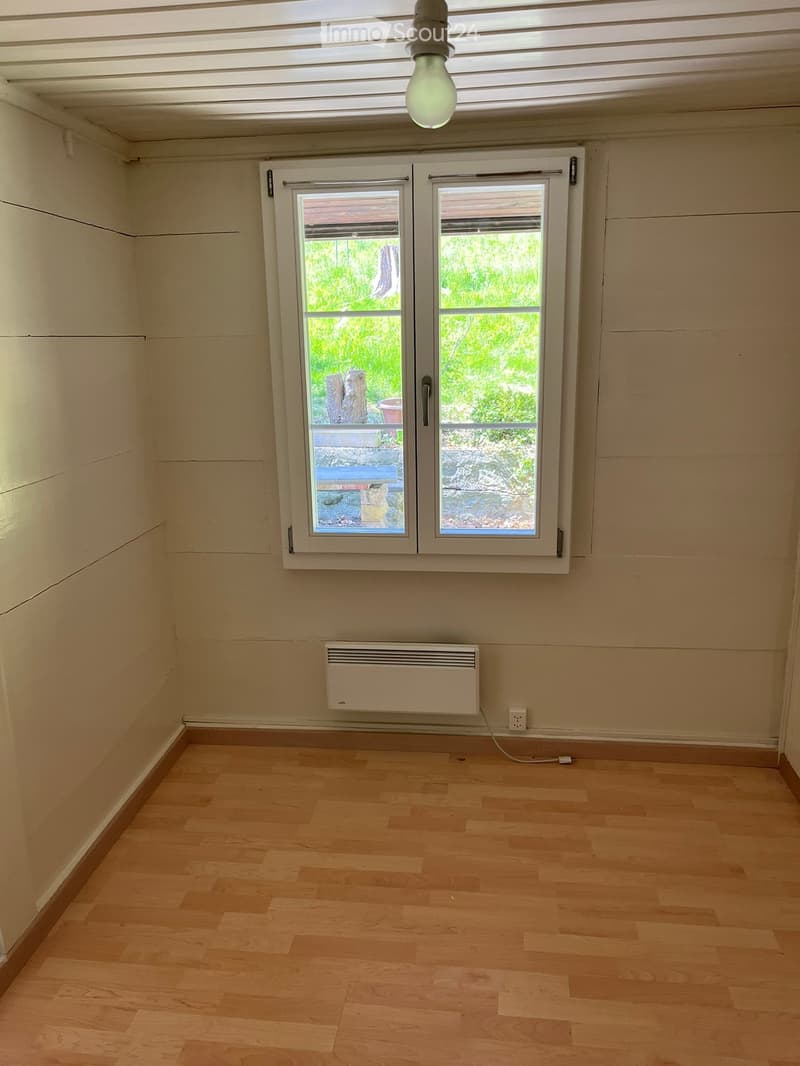Empty room with light wooden flooring, white walls, a window with a view of greenery, and a white radiator on the wall.