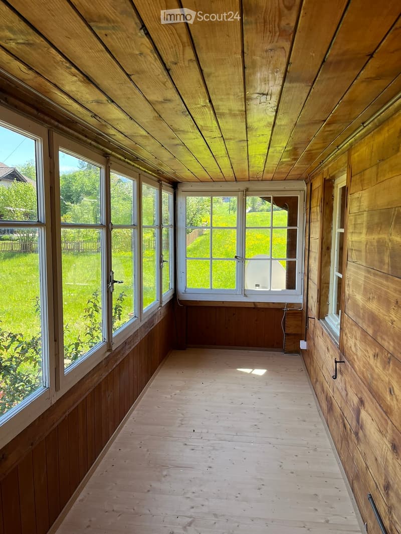 Empty wooden-framed room with large windows and a wooden floor, showing natural light and an outdoor view.