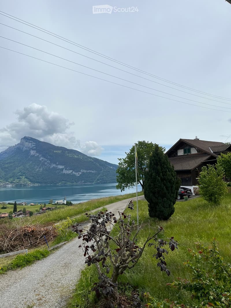 A scenic outdoor view featuring a house on a hillside, a lake in the background, and a mountain range. The foreground includes a gravel path and some greenery.