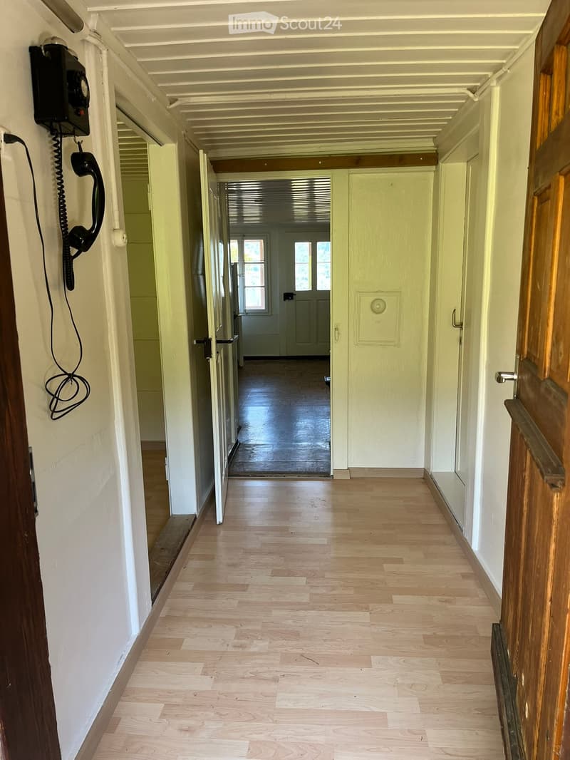 A long, narrow hallway with light wooden flooring, white walls, and several doors on both sides. A black telephone is mounted on the left wall, and the ceiling has a simple, white, paneled design.