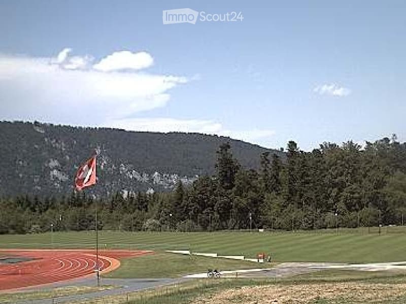 Outdoor track and field area with a running track, grassy field, and a flagpole with a flag, surrounded by trees and hills.