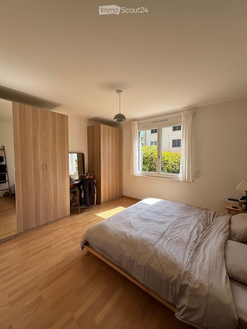 A modern, well-lit bedroom with wooden flooring, a large bed with grey bedding, a wardrobe, a desk with a chair, and a window with white curtains.
