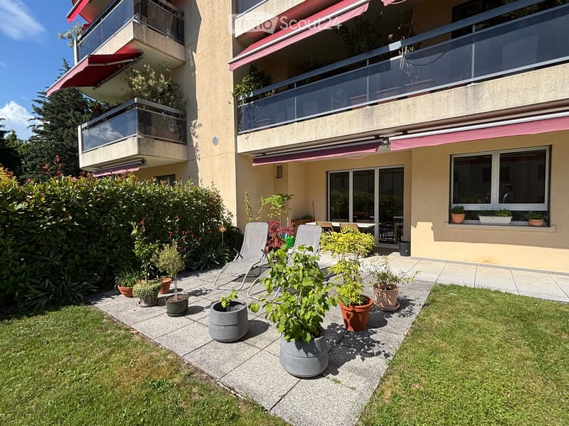 Garden area with potted plants, lounge chairs, and a small patio near a building with balconies and large windows.