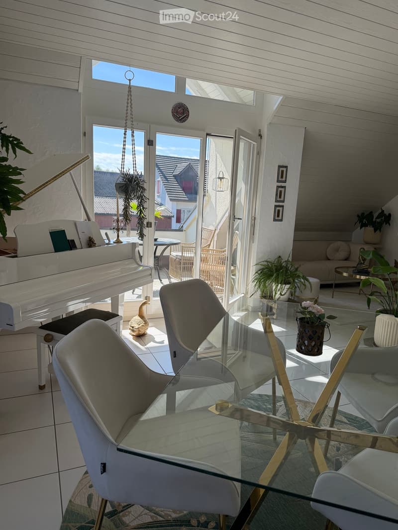 Bright living room with white walls, large windows, and a glass table surrounded by modern chairs. Features a white piano, decorative vase with flowers, and potted plants.