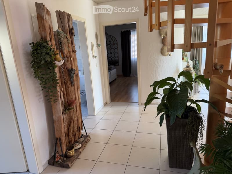 Hallway with tiled floor, wooden decorative piece on left, large potted plant on right, open doorway leading to another room.