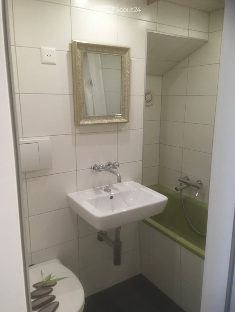 Modern bathroom with white tiled walls, a white sink with a chrome faucet, a decorative mirror, a toilet, and a bathtub with a green accent.