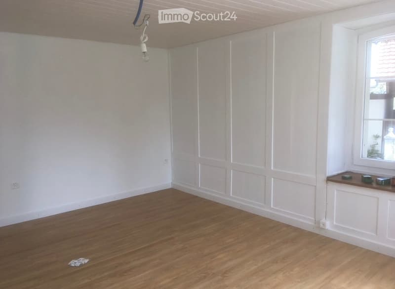 Empty room with wooden flooring, white paneled walls, and a single window with a small shelf holding a few items.
