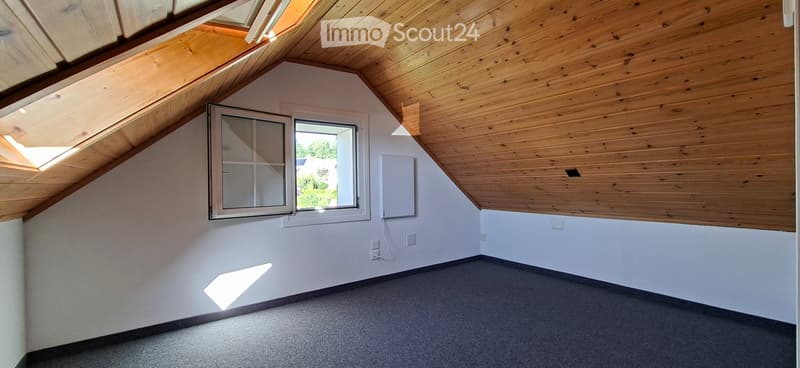 Empty attic space with wooden slanted ceilings, white walls, and a large window letting in natural light.