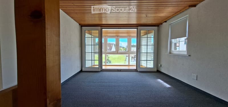 Empty room with grey carpet, light wooden ceiling, large sliding glass doors leading to a patio, and a window with blinds on the right side.
