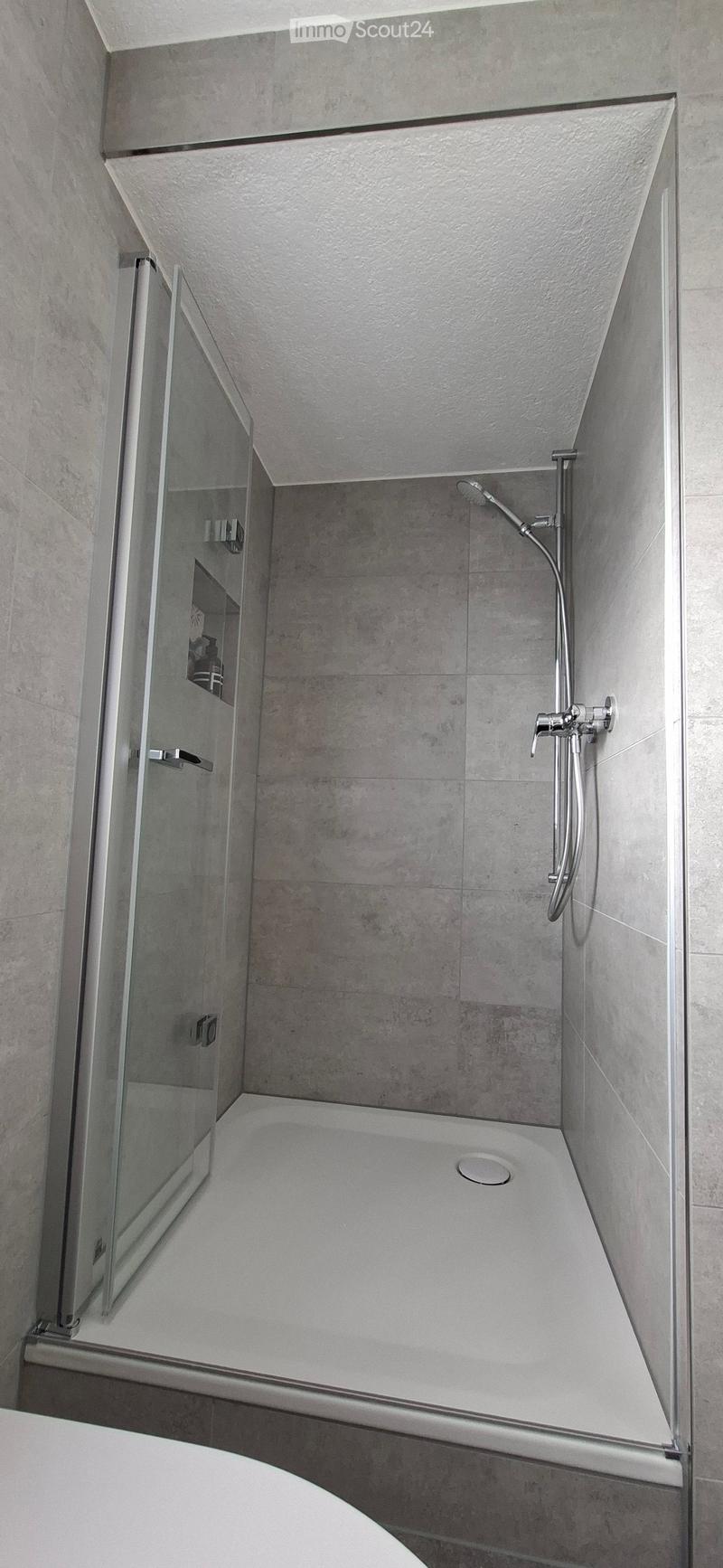 Modern shower stall with a glass enclosure, a handheld showerhead, and a built-in seat. The walls and floor are tiled in a neutral gray color.