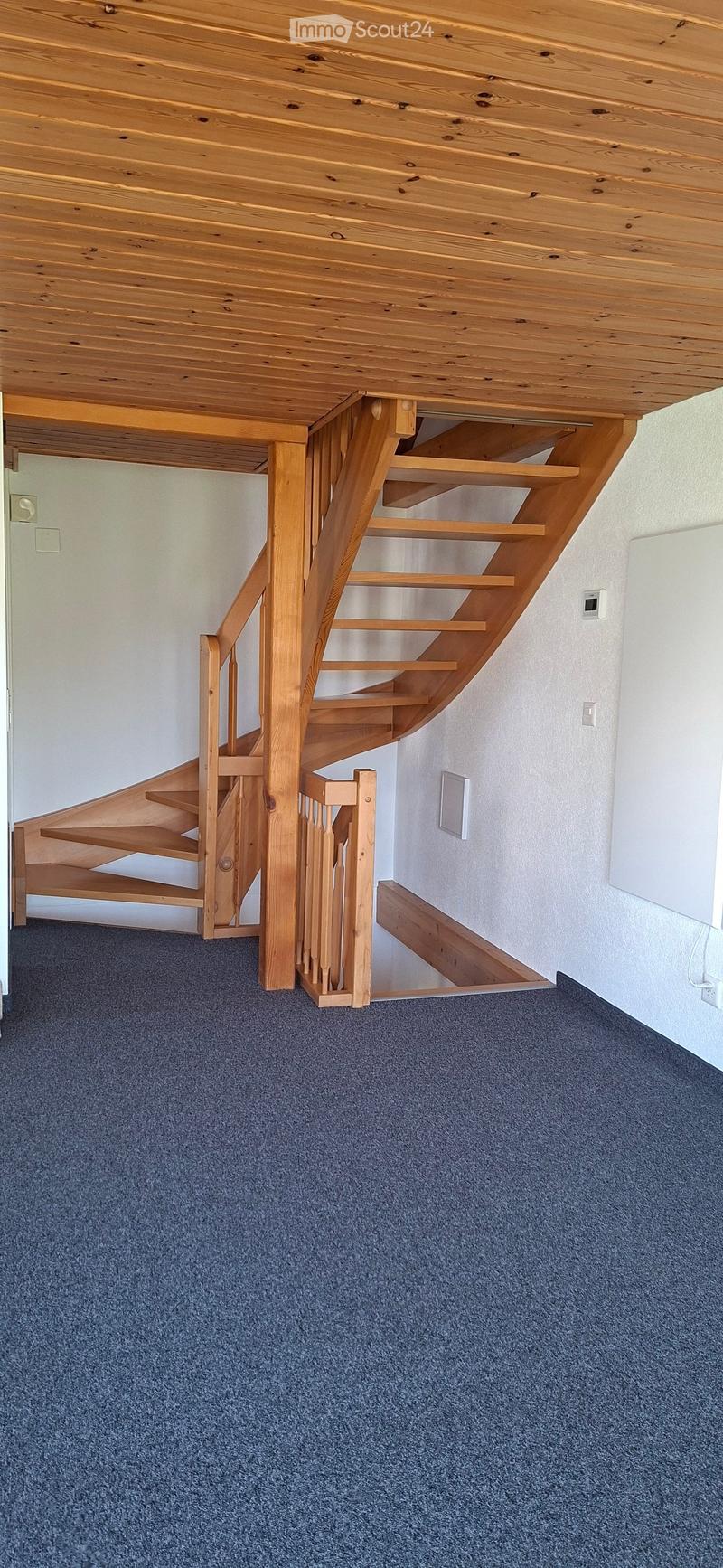 Wooden staircase with open wooden railings and steps, leading up to a wooden ceiling, with blue carpeted floor and white walls.