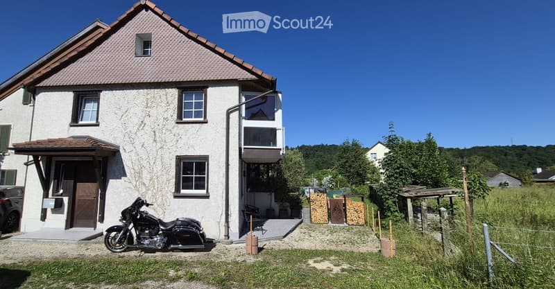 Two-story house with white exterior, multiple windows, and a balcony on the upper floor. A motorcycle is parked in front, and there is a small garden area with a wooden structure and stacked firewood.