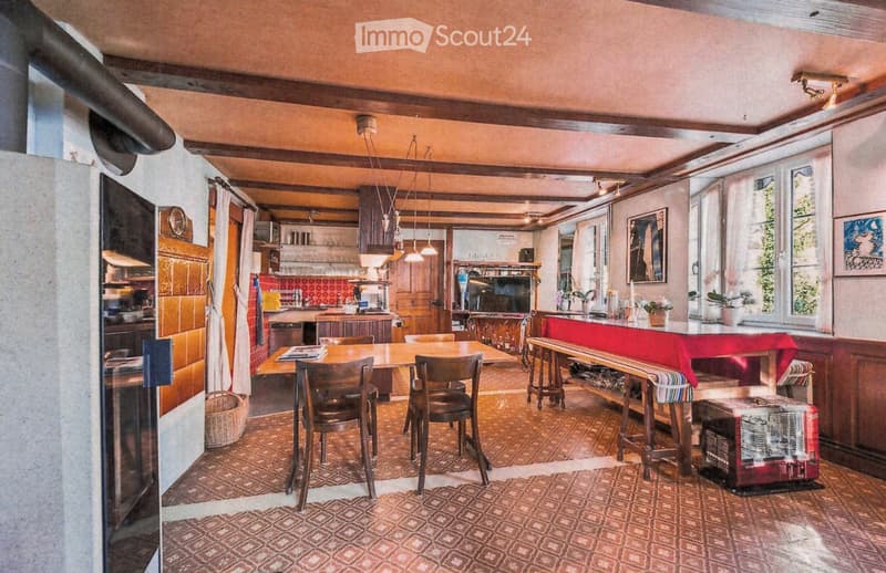 A cozy, well-furnished room with a dining area and an open kitchen. The space features wooden furniture, including a dining table with chairs, a bench, and a stool. A television is mounted on the wall, and the kitchen area includes cabinets and a counter.