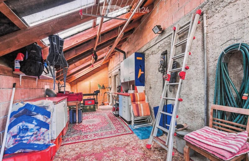 The image shows an attic or storage room with wooden beams and exposed rafters. Visible objects include a ladder, a chair, a stool, a cabinet, a table, and a backpack. The room has a rustic feel with wooden walls and a patterned rug on the floor.