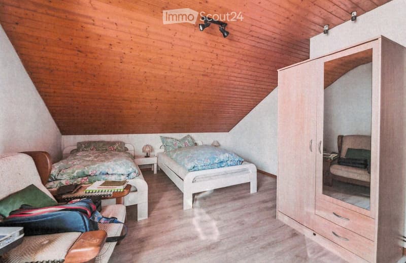 Cozy attic bedroom with wooden walls and slanted ceilings, featuring two single beds, a wardrobe with a mirror, a small table with a lamp, and a comfortable armchair.