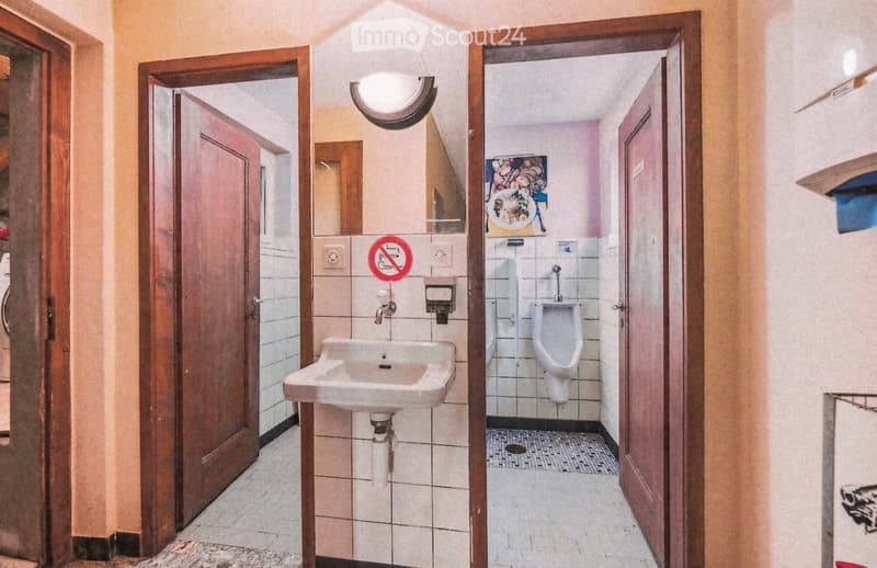 Public restroom with a sink, urinal, and a no-smoking sign. The area features tiled walls and floors, wooden doors, and a light fixture above the sink.