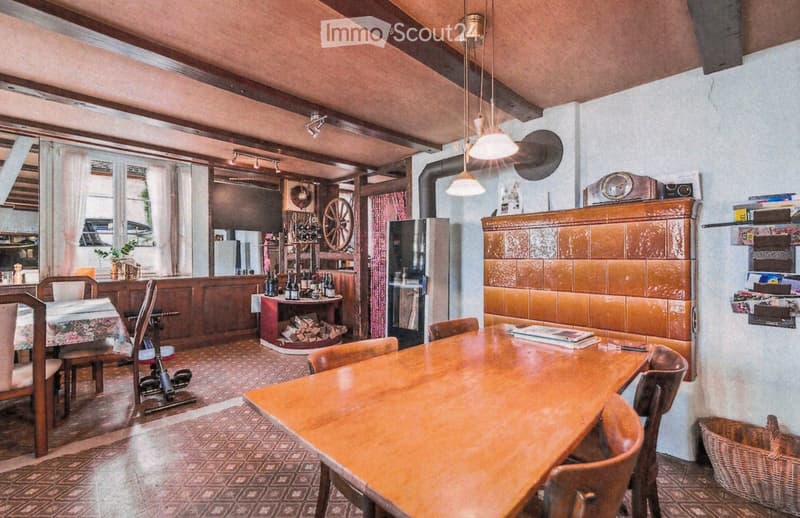 A cozy, rustic dining area with wooden furniture, a large wooden table surrounded by chairs, a wine rack, and a refrigerator in the background. The room features exposed wooden beams, warm lighting, and a mix of traditional and modern decor.