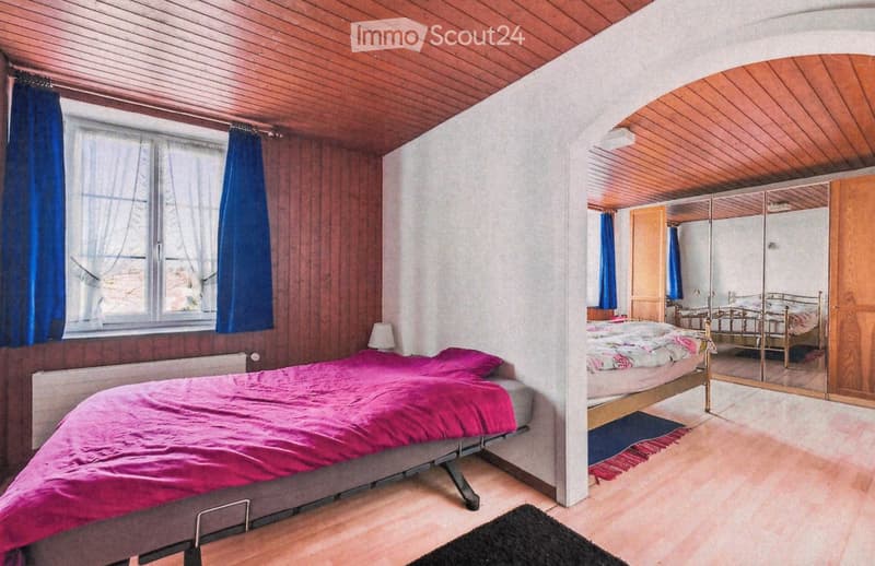 Cozy bedroom with wooden walls and ceiling, featuring a bed with pink bedding, a mirror, a wardrobe, and a table lamp.