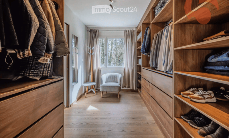 A well-organized walk-in closet with wooden shelves and drawers, a small seating area featuring a grey armchair and a round stool, and a coat rack with jackets and bags.