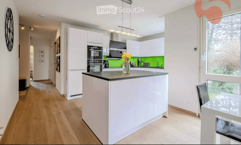 Modern kitchen with white cabinets, a central island, stainless steel appliances, and a green accent wall. Features a dining area with a glass table and black chairs.
