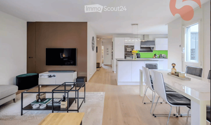 Modern open-plan living and dining area with a sofa, coffee table, TV, and a dining table with chairs. The kitchen features white cabinets, a kitchen island, and various appliances.