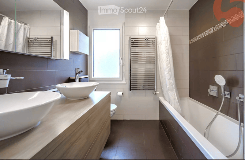 Modern bathroom with a large bathtub, a separate sink, a mirror, a towel, a radiator, and a toilet. The room features dark tiles and a wooden vanity.