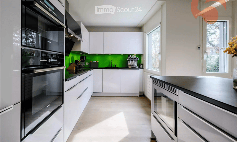 Modern kitchen with white cabinets, stainless steel appliances, and a green backsplash. Features an oven, fridge, sink, and coffee machine.