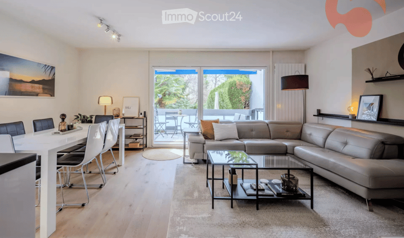 Modern living and dining area with a grey sectional sofa, glass coffee table, white dining table with chairs, and large windows opening to a balcony.