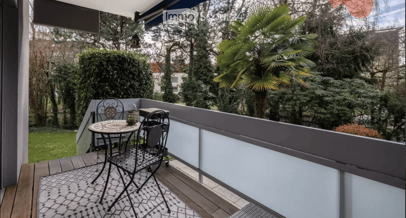 Balcony with wooden decking, modern railing, small round table with two chairs, and a lantern on the table. The view includes greenery and trees.