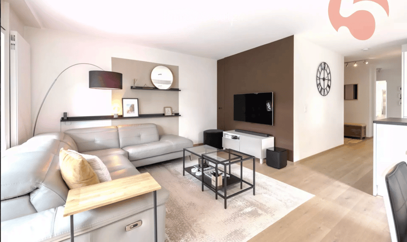 Modern living room with a grey sectional sofa, a glass coffee table, a floor lamp, a wall-mounted TV, a round mirror, and decorative shelves.