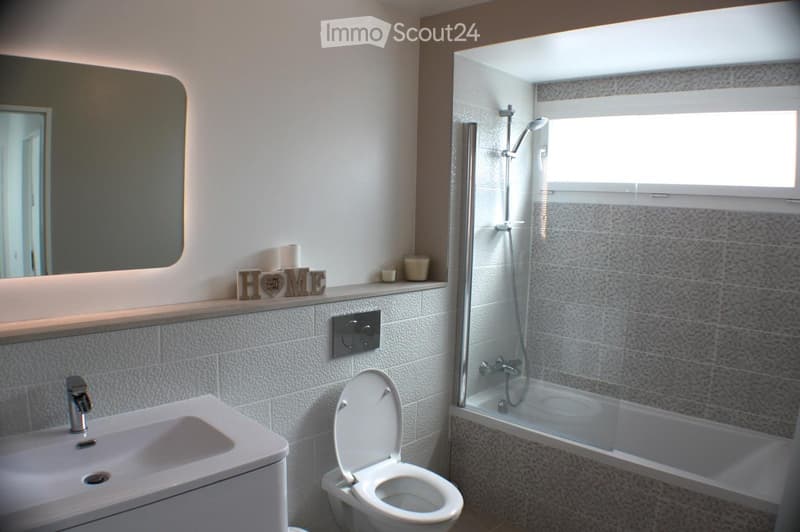 Modern bathroom with a white sink, toilet, shower, and a large mirror above the sink. The shower has a glass enclosure and a window above it, allowing natural light to enter.