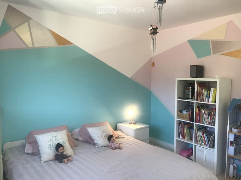 A cozy bedroom with a slanted ceiling, a bed with pillows and a blanket, a nightstand with a lamp, a bookshelf filled with books and a speaker, and decorative items including a wind chime and toys.