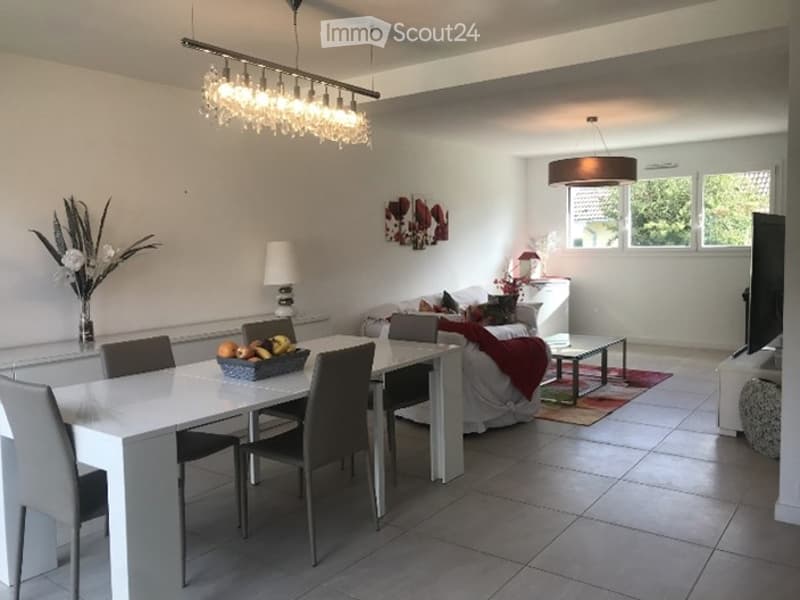 Modern, open-plan living and dining area with white walls, grey tiled flooring, and contemporary furniture including a sofa, dining table with chairs, coffee table, and decorative lamps.